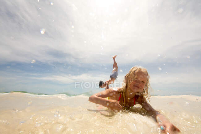 Girl lying on beach in shallow water, brother doing handstand in background — Stock Photo