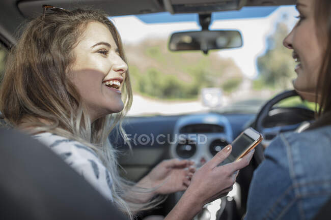 Mujer joven en coche con una amiga, sosteniendo el teléfono inteligente - foto de stock