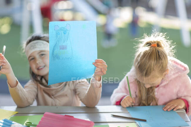 Zwei junge Mädchen, im Freien, zeichnen, junges Mädchen hält Kunstwerk hoch — Stockfoto