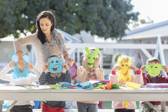 Metà donna adulta aiutare i bambini con attività artigianale, i bambini indossano maschere di carta — Foto stock