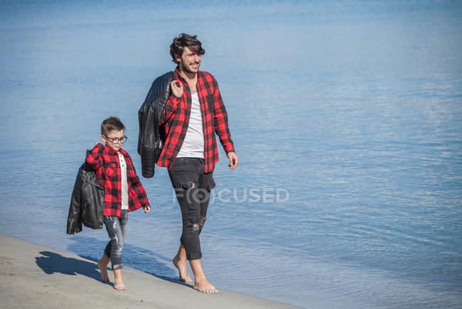 Vater und Sohn laufen am Strand entlang und tragen Jacken über der Schulter — Stockfoto