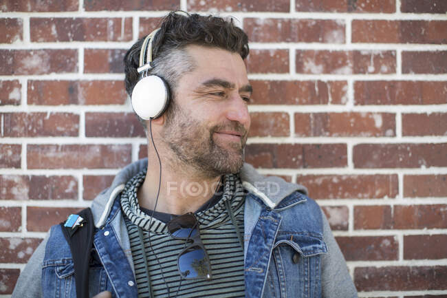 Hombre adulto medio apoyado contra la pared, usando auriculares, mirando hacia otro lado, sonriendo - foto de stock