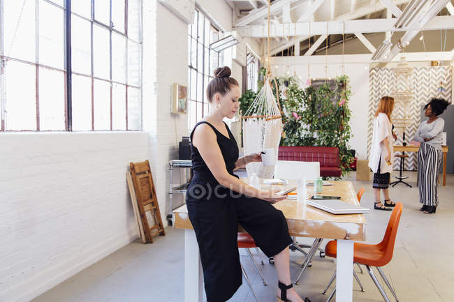 Woman sitting on desk in industrial office building and texting on smartphone — Stock Photo