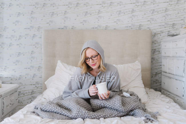 Young woman wrapped in blanket sitting on bed with cup of coffee — Stock Photo