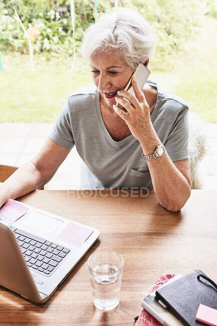 Mujer mayor sentada en la mesa con portátil delante de ella, usando un teléfono inteligente - foto de stock