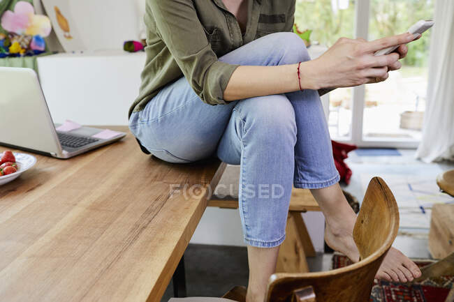 Mid section of woman sitting on kitchen table and using smartphone, low section — Stock Photo
