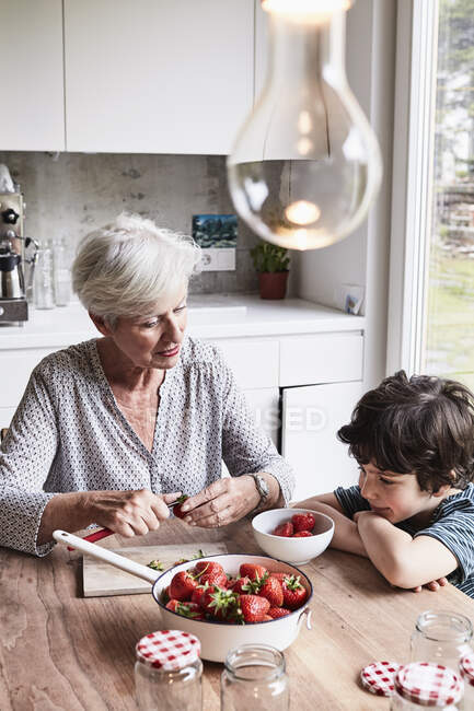 Großmutter sitzt am Küchentisch, bereitet Erdbeeren zu, Enkel sitzt neben ihr und schaut zu — Stockfoto