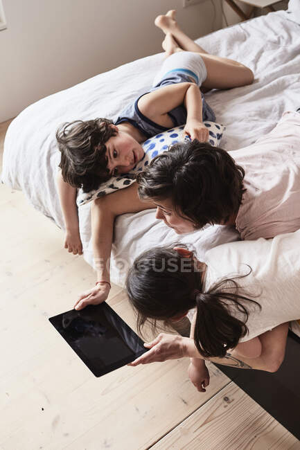Мати, син і дочка лежать на ліжку, використовуючи цифровий планшет, підвищений вид — стокове фото