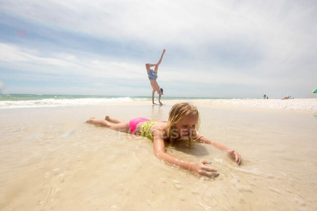 Girl lying on beach in shallow water, brother doing handstand in background — Stock Photo