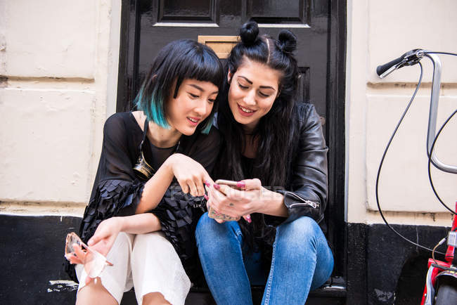 Две молодые стильные женщины со смартфоном сидят на пороге — стоковое фото