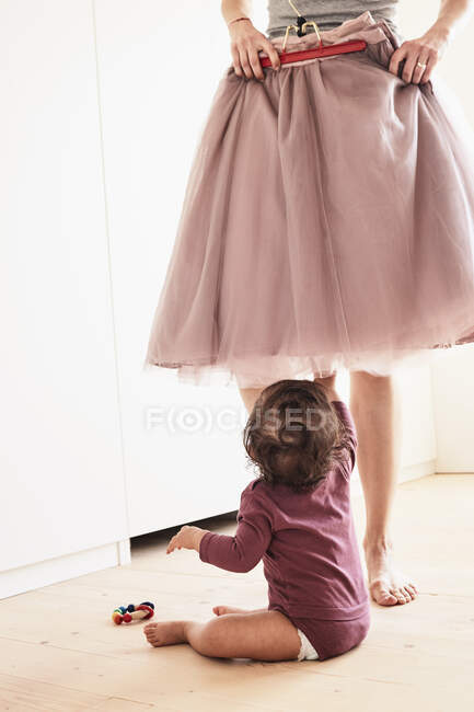 Mother holding skirt up against her, baby girl sitting on floor watching her, low section — Stock Photo