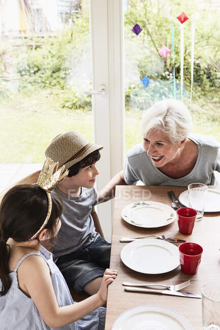 Бабушка сидит за кухонным столом с внуком и внучкой, улыбается — стоковое фото