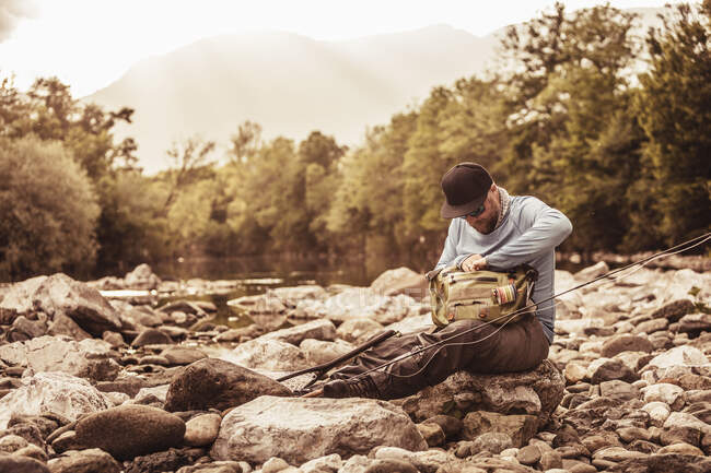 Fisherman sitting on river rocks searching fishing bag, Mozirje, Brezovica, Slovenia — Stock Photo