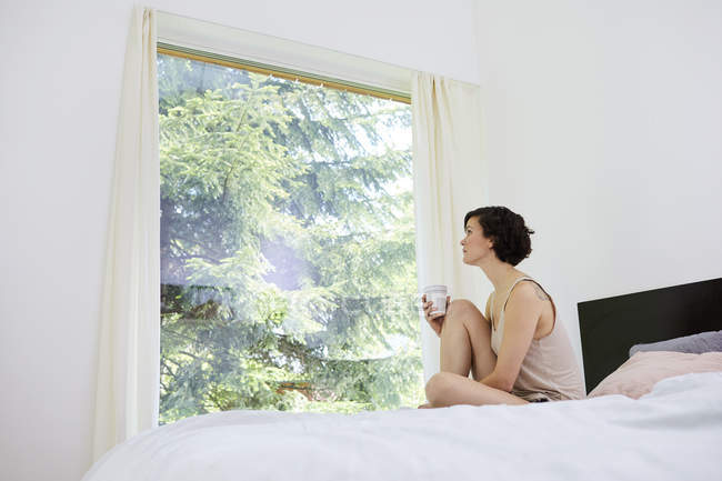 Взрослая женщина с кофейной чашкой сидит на кровати и смотрит в окно — стоковое фото
