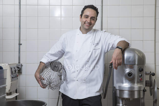 Портрет шеф-повара на коммерческой кухне, держащего венчик, смотрящего на камеру улыбающегося — стоковое фото
