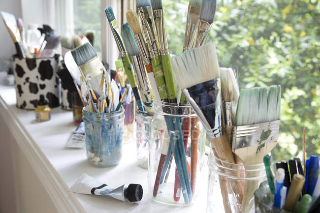 Fila de frascos con variedad de pinceles para artistas en el alféizar de la ventana del estudio de artistas - foto de stock