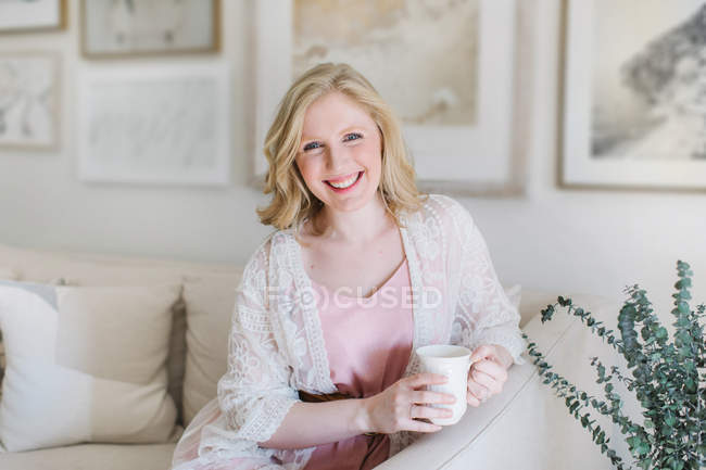 Jovem mulher sentada no sofá e segurando caneca de café — Fotografia de Stock