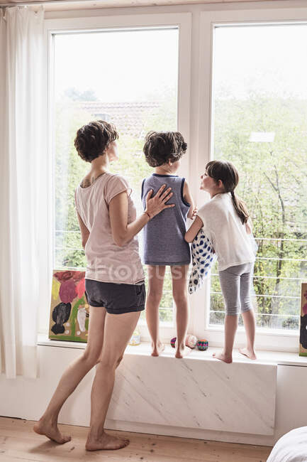 Madre, hijo e hija en casa, mirando por la ventana, vista trasera - foto de stock