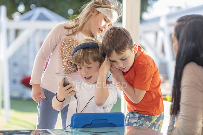 Tres niños pequeños, usando smartphone, escuchando a través de auriculares - foto de stock