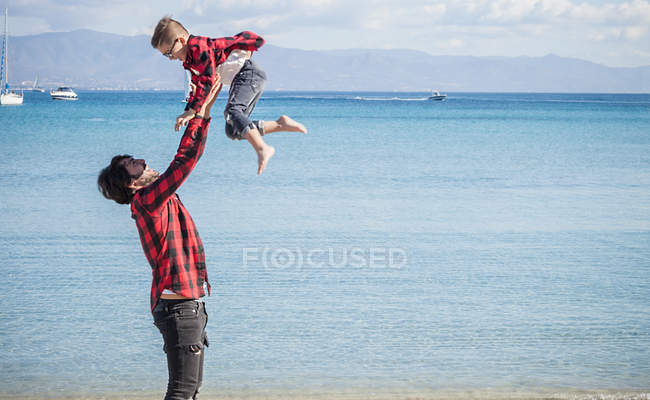 Padre levantando hijo en el aire en la playa - foto de stock