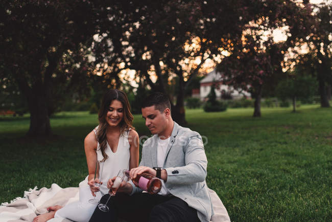 Romántica pareja joven vertiendo champán rosa en el parque al atardecer - foto de stock