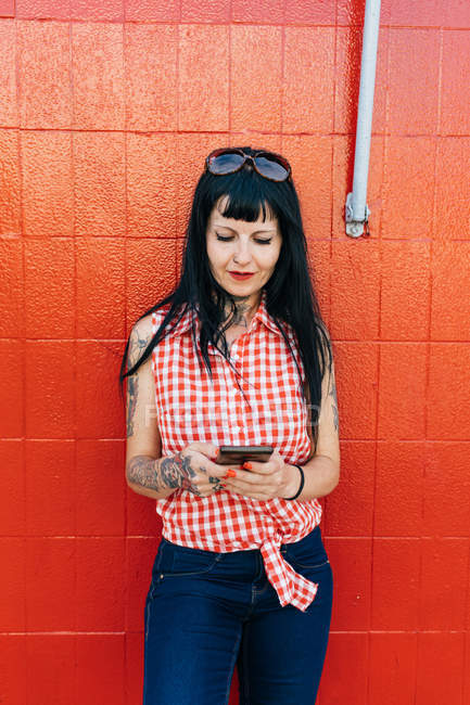 Mujer madura hipster apoyado contra la pared roja mirando el teléfono inteligente - foto de stock