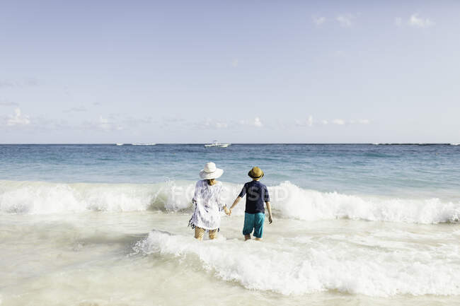 Мать и сын, держась за руки, стоя в серфинге на пляже, вид сзади — стоковое фото