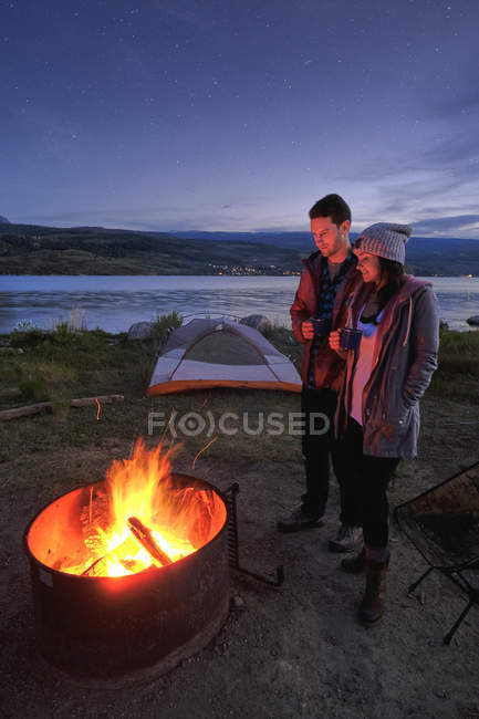 Paar steht in der Abenddämmerung mit heißen Getränken am Lagerfeuer — Stockfoto