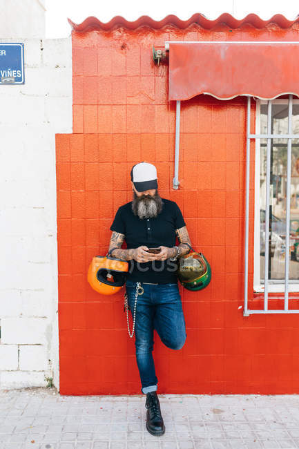 Male hipster leaning against red wall looking at smartphone — Stock Photo