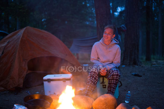 Adolescente sentado junto al fuego del campamento en el camping - foto de stock