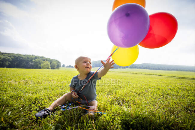 Baby boy sitting in rural field with bunch of colourful balloons — Stock Photo