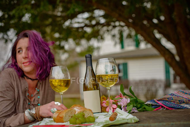 Retrato de mujer sentada a la mesa con botella de vino, vasos y comida al aire libre - foto de stock