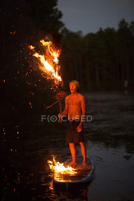 Giovane uomo che tiene ramo di albero in fiamme quando in piedi vicino all'acqua al tramonto — Foto stock
