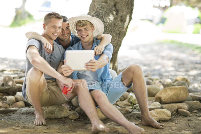 Троє молодих друзів-чоловіків сидять на каменях, дивлячись на цифровий планшет — стокове фото