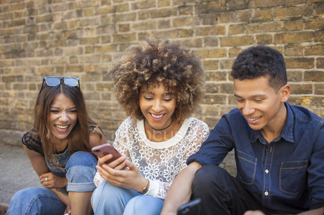 Tres amigos sonrientes mirando el teléfono inteligente sentado en la calle - foto de stock