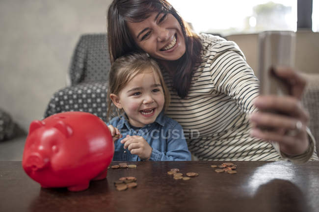 Mature woman taking selfie with daughter and piggy bank — Stock Photo