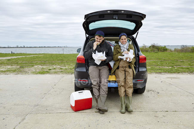 Young couple sitting in open car trunk and eating food — Stock Photo