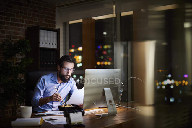 Молодой бизнесмен читает бумажки и ест еду на вынос в офисе ночью — стоковое фото