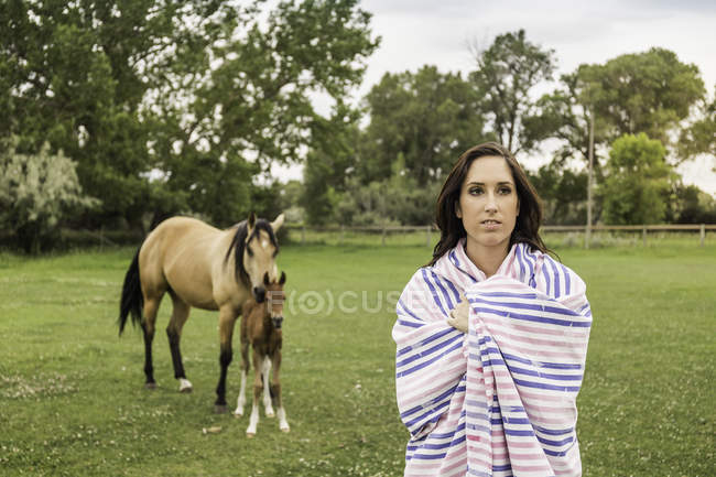 Portrait of young woman wrapped in blanket, horse and foal in background — Stock Photo