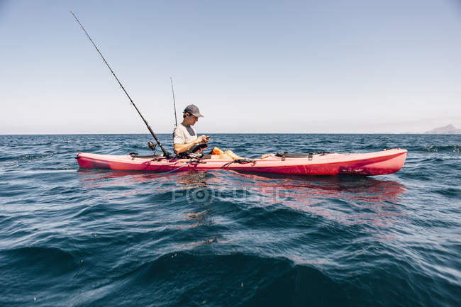 Junger männlicher Seekajakfahrer schaut beim Angeln aufs Smartphone, Santa Cruz Island, Kalifornien, USA — Stockfoto