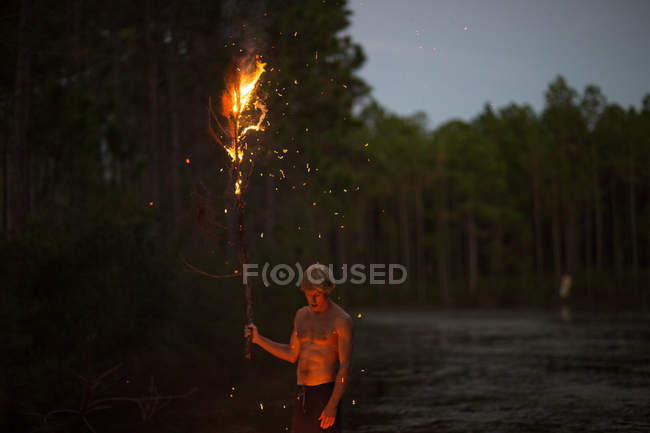 Young man holding burning tree branch when standing near water at dusk — Stock Photo