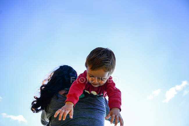 Vista de ángulo bajo del niño sobre el hombro de la madre contra el cielo azul - foto de stock