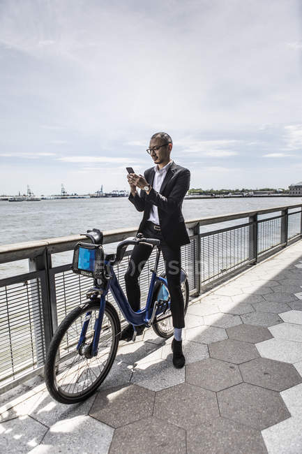Businessman on bicycle looking at smartphone along city river waterfront — Stock Photo