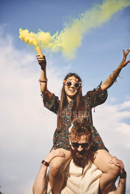 Young boho woman holding yellow smoke flare on boyfriend shoulders at festival — Stock Photo