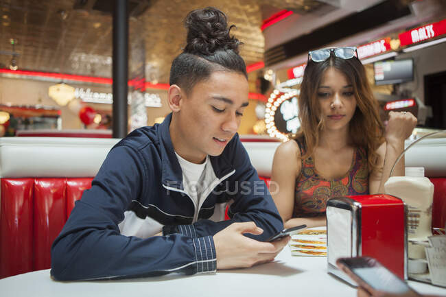 Молодая пара, сидящая в закусочной, молодой человек смотрит на смартфон, женщина со скучным выражением лица — стоковое фото