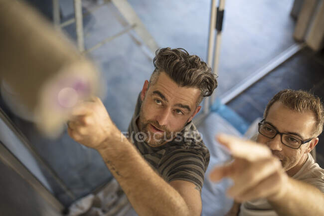 Mann bemalt Wand mit Farbroller, männlicher Freund zeigt auf Malfläche, erhöhter Blick — Stockfoto