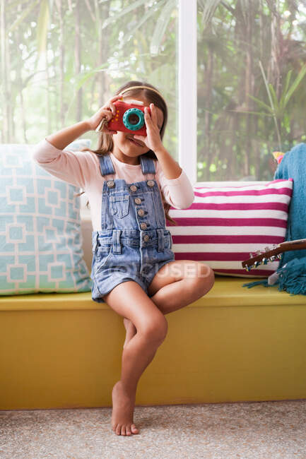 Retrato de niña en el asiento de la ventana fotografiando con cámara de juguete - foto de stock