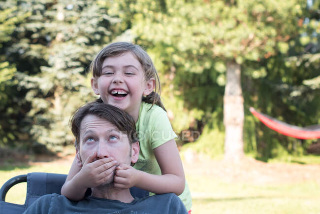 Padre e hija jugando al aire libre, chica joven cubriendo la boca del padre, riendo - foto de stock