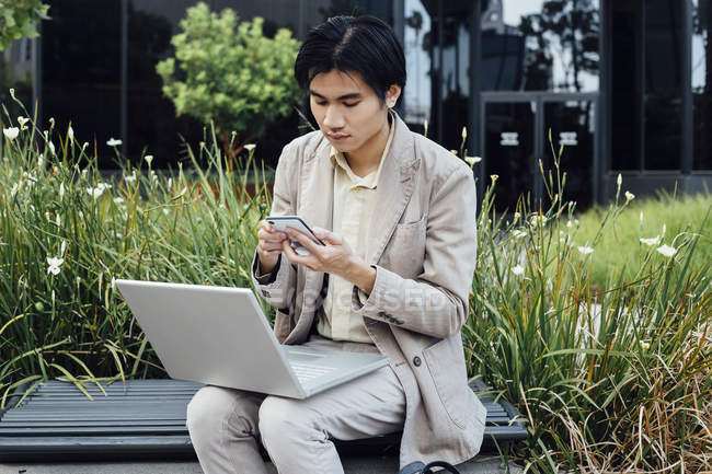 Hombre joven usando el ordenador portátil y el teléfono inteligente al aire libre - foto de stock