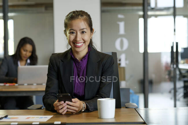 Retrato de mulheres de negócios sentadas na mesa, segurando smartphone, sorrindo — Fotografia de Stock